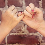 Photographie de deux mains qui croisent leurs doigts devant un mur de briques rouges.