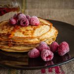 Photographie d'une assiette de pancakes, avec quelques framboises et du sirop d'érable.