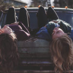 Photographie de deux femmes allongés sur le dos, sur le capot d'une voiture. Les cheveux longs qui glissent vers le sol.