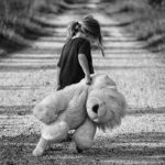 Photographie d'une petite fille triste qui marche sur un chemin de terre, traînant une grande peluche.