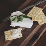 Photographie d'un plateau en bois, portant des crackers, un couteau et du fromage frais.