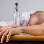 Cette image illustre un homme qui dort la tête sur la table, à côté d'une bouteille d'alcool vide.